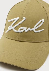 Beige beisbola cepure ar izšūtu baltu "Karl" tekstu, strukturētu kroni, izliekto malumu un teksturētu auduma apdari.