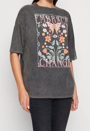 Person trägt ein graues übergroßes T-Shirt mit einem bunten Blumen- und Schmetterlingsdesign und dem Text „EMBRACE CHANGE“ auf der Vorderseite.