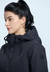 Giacca nera impermeabile con colletto alto, cappuccio regolabile con cordino e una patch con logo sulla spalla. Texture liscia e opaca.