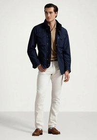 Άνδρας με ναυτικό σακάκι, μπεζ πουκάμισο, λευκά παντελόνια και καφέ loafers που στέκεται σε απλό φόντο.