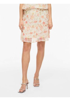 Žena nosi svetlú kvetinovú sukňu s volánikmi, v kombinácii so striebornými sandálmi na vysokom opätku, stojaca na bielom pozadí.