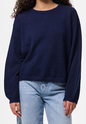 Femme portant un pull bleu marine à manches longues et un jean denim bleu clair, debout devant un fond blanc uni.