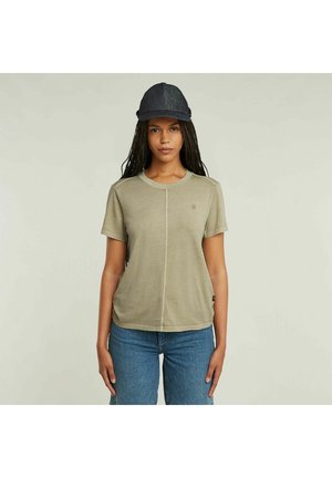 Donna con lunghi capelli trecciati che indossa una T-shirt khaki a maniche corte, jeans blu e un berretto di denim scuro su uno sfondo semplice.