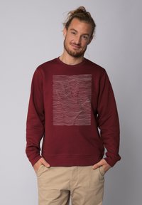 Sweat à col rond bordeaux avec un graphique texturé en blanc représentant une main au milieu de lignes ondulées, avec des poignets côtelés.