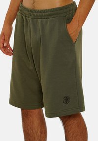 Olivgrüne Shorts aus weichem Material, mit elastischem Bund, Seitentaschen und einem dezenten schwarzen Logo auf dem Unterschenkel.