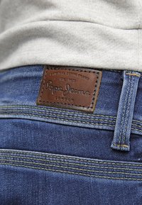 Blå denim jeans med ett brunt läderpatch som har "Pepe Jeans"-logotypen. Gula sömmar och en standard knappstängning.