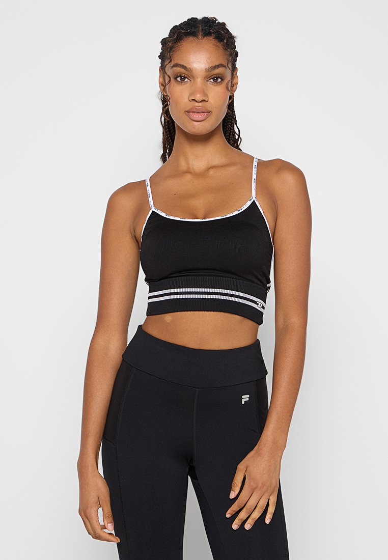Jeune femme avec des cheveux tressés portant un crop top noir à fines bretelles blanches et un legging noir taille haute avec un logo sur la cuisse gauche.