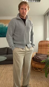 Siv krojeni pulover na zadrgo z visokim ovratnikom, kombiniran s svetlim bež hlačami. Sproščen kroj, prikazan v dobro osvetljenem notranjem okolju.