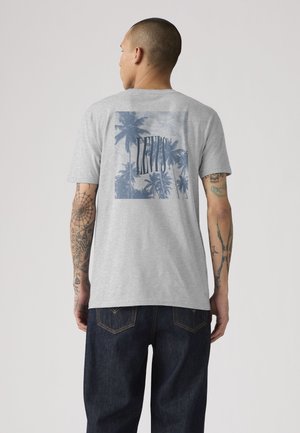 Homme avec des tatouages portant un t-shirt gris avec des palmiers bleus et le texte "LEVI'S", associé à un jean bleu foncé, vu de dos.