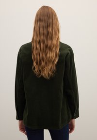 Dark green corduroy shirt, long-sleeved with a straight cut. Textured fabric, visible ribbing, and buttoned cuffs. Worn with navy jeans.