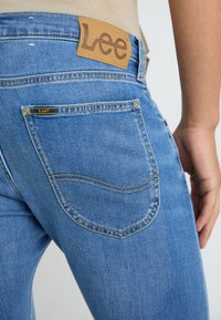 Jean en denim bleu clair avec un design distinct des poches arrière, comportant une étiquette en cuir avec "Lee" en marron et des accents de couture jaune.
