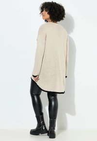 Crème breigo veredeld met zwarte bies, lange mouwen en een iets asymmetrische zoom, gecombineerd met glanzende zwarte leggings en enkellaarsjes.