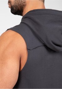 Mouwloze capuchonsweater van donkergrijs stof. Kenmerkt zich door een ruwe rand bij de schouder en een ontspannen pasvorm. Soepele, zachte textuur.