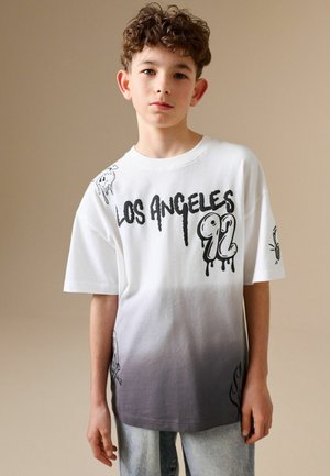 Chłopiec o kręconych włosach, ubrany w białą-szarą koszulkę ombre z napisem „Los Angeles 92” i czarnymi wzorami graficznymi, stojący na jednolitym tle.