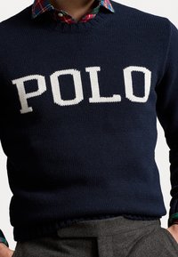 Mörkblå stickad tröja med stora vita bokstäver "POLO" på framsidan, rund halsringning och ribbat fåll.