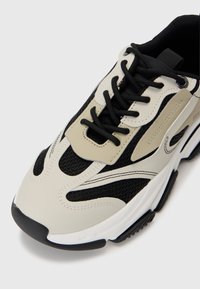 Beige and black sneaker featuring mesh panels, thick black laces, a distinctive rubber sole, and subtle wave patterns on the upper.