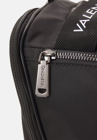 Saco de nylon preto com um puxador de zíper prateado com a gravação "VALENTINO", alça texturizada e detalhes de costura em contraste.