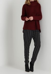 Bordowy sweter z prążkowanej dzianiny z wysokim kołnierzem w połączeniu z czarnymi spodniami w prążki i botkami za kostkę, tworząc warstwowy wygląd.