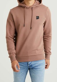 Mikina v rezavé barvě s rybinovou strukturou, se stahovací kapucí a malou černou logem na hrudi. Má dlouhé rukávy a přiléhavý lem.