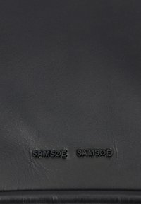 Czarna skórzana powierzchnia z tłoczonym logo "SAMHØE SAMHØE". Gładka faktura z delikatnym połyskiem i minimalnymi detalami szwów.