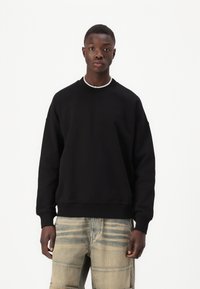 Svart hettegenser med rund hals og avslappet passform, med brodert logo på brystet. Kombinert med lyst, mønstret denimjeans.
