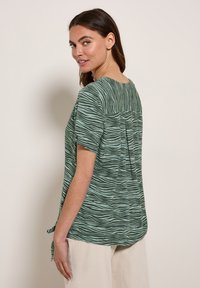 Groene shirt met een gestructureerd, golfachtig patroon, korte mouwen en een detail met plooien aan de achterkant. De zoom is afgerond en iets langer aan de achterkant.