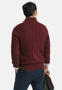 Burgunderfarbener Kabelstrickpullover mit hohem Kragen und geripptem Saum, mit einem strukturierten Muster und langen Ärmeln, getragen über einem gestreiften Hemd.