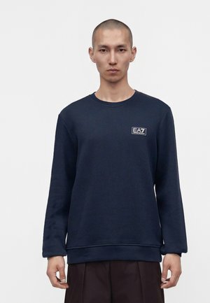 Homme aux cheveux courts portant un sweat-shirt à manches longues bleu marine foncé avec un logo EA7 Emporio Armani sur la poitrine.