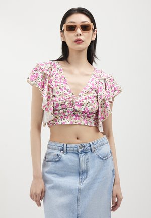 Donna con occhiali da sole rettangolari beige, un top corto arricciato floreale rosa e una gonna di denim alta a vita leggera blu chiaro su sfondo bianco.