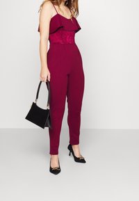 Bordový overal s volánkovými ramínky a krajkovým korzetem, doplněný černými lodičkami na podpatku a malou černou kabelkou.