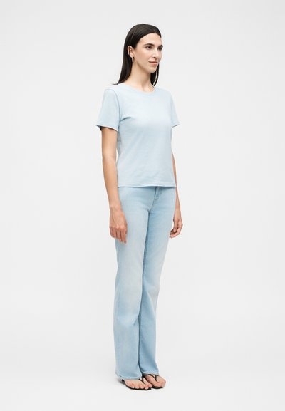 Γυναίκα που στέκεται, φοράει ανοιχτό μπλε κοντομάνικο πουκάμισο, ανοιχτό μπλε τζιν και μαύρα σαγιονάρες, πάνω σε λευκό φόντο.