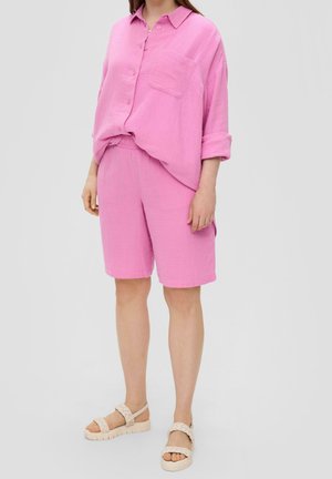 Žena v růžové volné košili na knoflíky s vyhrnutými rukávy a odpovídajících růžových šortkách do kolen, v kombinaci se světle krémovými sandály s pletenými řemínky.