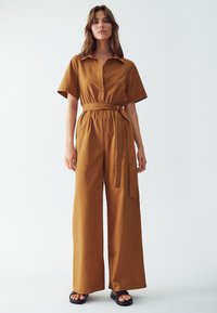 Hnědý overal s páskem, límečkem, krátkým rukávem a volným střihem. Vyroben z lehké látky s hladkou texturou. Černé sandály.