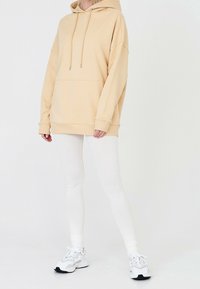 Béžová oversized mikina s klokáním kapsou a šňůrkami, kombinovaná s bílými legínami a světle šedými teniskami. Hladká textura látky.