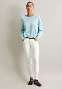 Hellblauer Strickpullover mit strukturiertem Muster, Rundhalsausschnitt und gerippten Bündchen, kombiniert mit weißen enganliegenden Hosen und schwarzen geflochtenen Sandalen.
