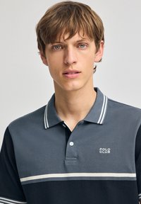 Pikétröja i mörkgrå och marinblå, med ett tvåfärgat rand över bröstet, en polokrage och en logotyp på vänster sida.