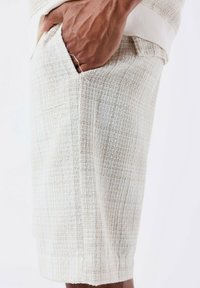 Shorts beige clair en tissu tissé texturé. Dotés d'une poche latérale et d'un logo discret. Ourlés aux genoux avec une coupe décontractée.