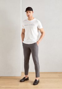 White t-shirt with grey text, grey slim-fit trousers, and black loafers. Model stands with hands in pockets against a neutral background.