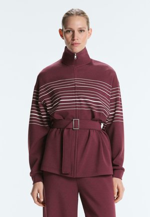 Žena nosi bordo jaknu na patentni zatvarač s horizontalnim bijelim prugama i remenom u struku, uparenu s odgovarajućim bordo hlačama.