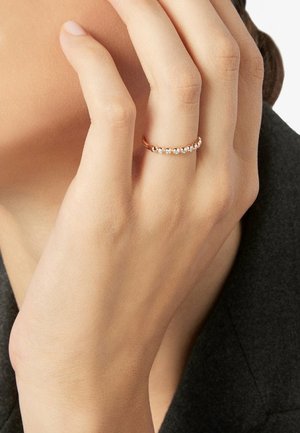 Hand mit roséfarbenem Goldring mit einer Reihe kleiner, klarer Edelsteine, die nahe der Wange ruht und ein dunkles Kleidungsstück trägt.