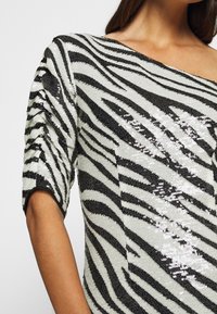 Haut à imprimé zèbre orné de sequins, avec un décolleté asymétrique et des manches courtes froncées. Principalement blanc avec des accents noirs.