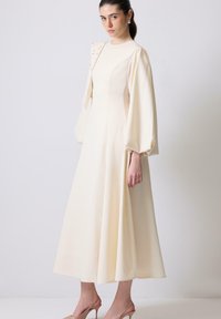 Nő egy krém színű midi ruhában, amely puffos hosszú ujju, aszimmetrikus szegecses részlettel az egyik vállán, és bézs visszahúzható sarkú cipőben áll egy fehér fal előtt.