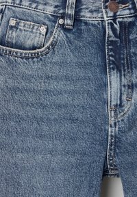 Jean en denim bleu en gros plan montrant la poche avant, la passant de ceinture, le bouton avec gravure de la marque, et les coutures détaillées sur un tissu texturé.