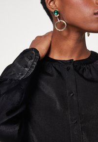 Blouse en satin noir avec un col rond, des fermetures à boutons et des poignets à volants. Comprend des boucles d'oreilles remarquables avec des pierres vertes et des accents circulaires.