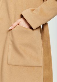 Beige coat with a soft, textured fabric. It features a large patch pocket on the front and a wide cuff on the sleeve.