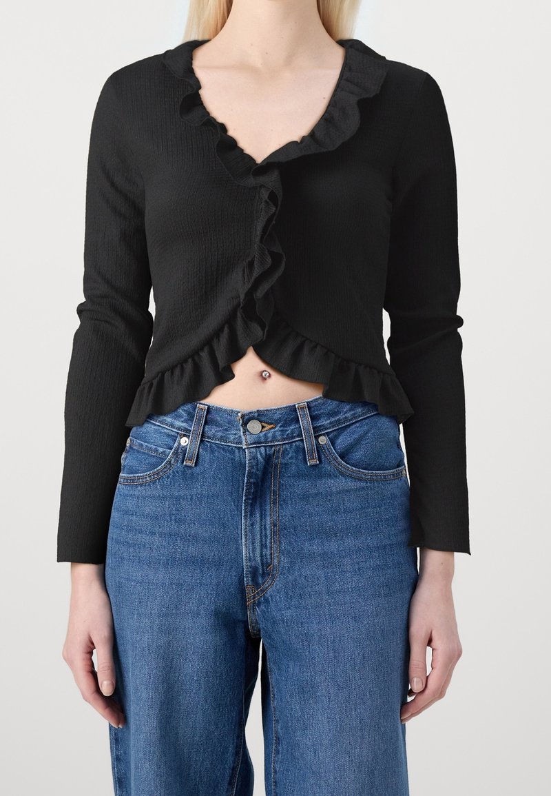 Femme portant un haut noir à manches longues avec des volants et un jean bleu, montrant un piercing au nombril.
