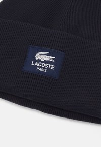 Beanie blu navy in maglia con un design a coste testurizzate e un patch rettangolare del logo Lacoste in bianco su uno sfondo blu scuro.