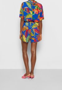 Conjunto colorido de camisa de manga corta y pantalones cortos con patrones de hojas tropicales y flores sobre un fondo azul, combinado con sandalias de tacón rosas.