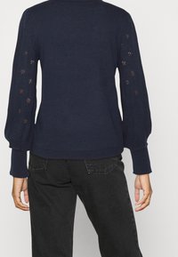 Mörkblå stickad tröja med puffärmar och mönster i öppen väv på ärmarna. Ribbad mudd och nederkant. Mörka jeans synliga nedanför.