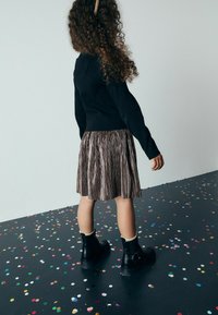 Top lungo a maniche lunghe nero a coste, gonnellina pieghettata bronzo metallico e stivaletti neri alla caviglia con fibbia. Confetti colorati su un pavimento scuro.